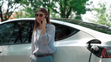 Doğal orman ya da milli parktaki araba yolculuğu sırasında elektrikli araba pili şarj ederken kadın telefonda konuşuyor. Tatil ve tatil boyunca çevre dostu seyahat. Yükselt