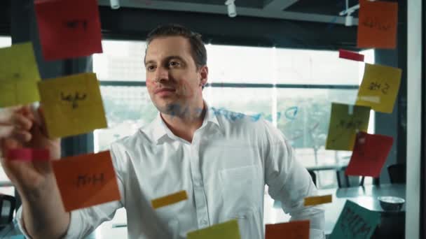 专业的男性领导者用贴纸写营销理念 白人商人在玻璃墙边用彩色粘贴贴纸思考营销活动 穿上衣服机械手 — 图库视频影像