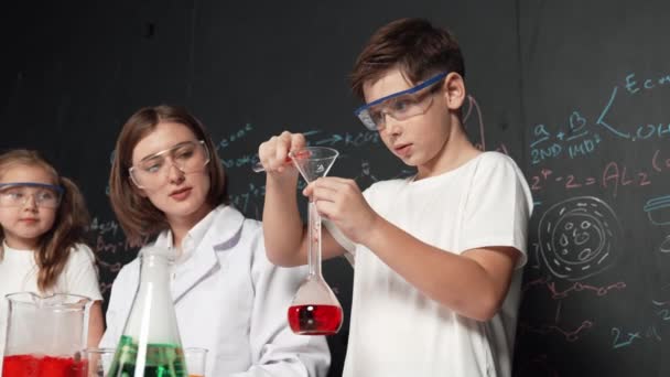 スマートな少年はビーカーに化学液を注ぎ 教師はStemサイエンスクラスでアドバイスをしています 学生は 実験室で化学理論を書かれたボードに立っている間 着色ソリューションを混合します エラー — ストック動画