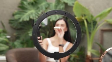 Dairesel halka ışığının merkezine yakınlaştır. Kadının güzellik ve kozmetik video içeriği için selfie çubuğu kullandığını göster. Doğal bahçenin huzurlu ortamında bir güzellik blogcusu. Blithe