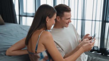 Genç çift, kredi kartıyla ödemek için internet üzerinden ödeme uygulaması ve dijital cüzdan kullanarak yatak odasında oturuyor. Mobil internet üzerinden alışveriş ve modern alımlar. Düzenle