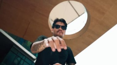 İspanyol dansçı alışveriş merkezinde dans ederken kameraya bakıyor. Çekici yakışıklı hippi ellerini alçak açılı kamerayla modern alışveriş merkezinde hip hop müziğine yöneltiyor. Açık hava sporu 2024. Sokak dansı. hiphop.