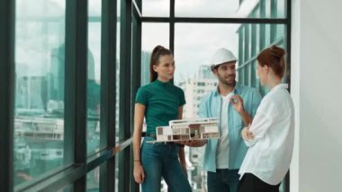 Proje yöneticisi ev modelini elinde tutarken, mimar mühendis takımı da ev tasarımı veya bina inşaatı hakkında fikir alışverişinde bulunuyor. Şehrin penceresinin yanında iç mimar duruyor. İzleme