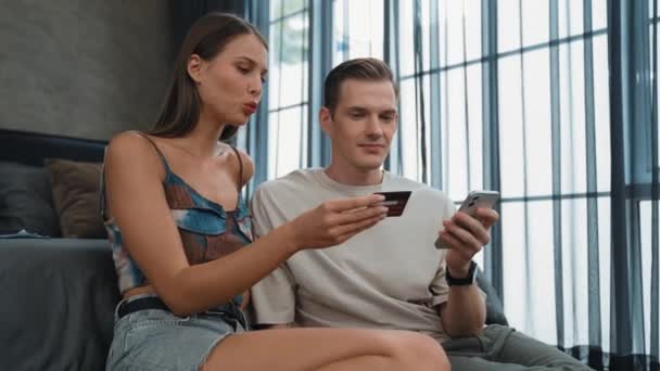 若いカップルは クレジットカードで支払うためにスマートフォンでオンライン決済アプリとデジタルウォレットを使用してホームベッドルームに座っています モバイルインターネットを介して商業ショッピングとモダンな購入 アディット — ストック動画
