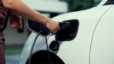 Kadın elektrikli arabasını akıllı ev şarj istasyonunda bulunan EV şarj cihazıyla şarj ediyor. Fütürist dijital batarya durumu hologramı. Sürdürülebilir güç için EV araba ve ev enerjisi altyapısı. İnceleyin