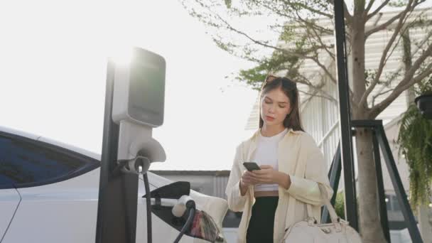 慢动作的年轻女性在城市购物中心停车场为电动车充电时 会在智能手机上检查电池的状况 现代女人用电动车购物 — 图库视频影像