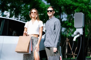 Genç çift yazın yeşil sürdürülebilir şehir bahçesinde EV elektrikli araba ile seyahat ederken, yeşil temiz yeniden şarj edilebilir elektrikli araç iç organları ile kentsel sürdürülebilirlik yaşam tarzını gösteriyor.