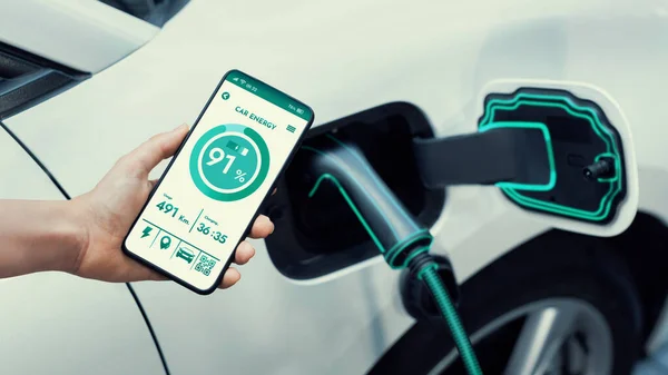 スマートフォンのディスプレイバッテリーステータスインターフェースをスマートEvモバイルアプリケーションで保持するビジネスウーマンと 駐車場の充電ステーションから電気を充電するEvカー Peruse — ストック写真