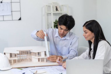 Profesyonel Asyalı erkek mimar ev modelinin uzunluğunu ölçmek için cetvel kullanırken, beyaz tenli genç meslektaşı ev modeliyle toplantı masasındaki verileri analiz etmek için dizüstü bilgisayar kullanıyor. Tertemiz..
