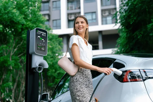 若い女性は夏に緑の持続可能な都市の屋外庭で充電するEv電気自動車で旅行します 電動Bev車両旅館のグリーンクリーン再充電可能エネルギーによる都市持続可能性ライフスタイル — ストック写真