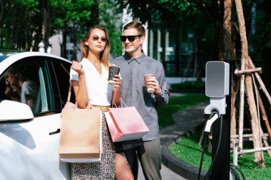 Genç çift yazın yeşil sürdürülebilir şehir bahçesinde EV elektrikli araba ile seyahat ederken, yeşil temiz yeniden şarj edilebilir elektrikli araç iç organları ile kentsel sürdürülebilirlik yaşam tarzını gösteriyor.