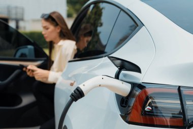 Genç bir kadın EV otomobil şarj istasyonu Green City Park 'ta elektrik ödemek için akıllı telefonu kullanıyor. Modern çevre ve EV araçlı sürdürülebilir şehir yaşam tarzı. Yararlı