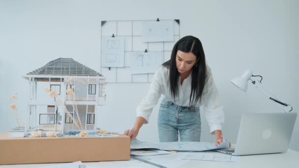 年轻美丽的高加索建筑师把蓝图摆到桌上 并与房屋模型进行比较 同时在现代办公室仔细检查带有条纹的蓝图的模型建设 纯洁无瑕 — 图库视频影像