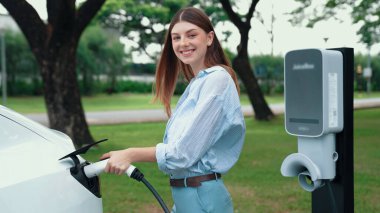 Yolculuk sırasında doğal orman ya da milli parktaki elektrikli araba için şarj olan genç bir kadın. Tatil ve tatil boyunca çevre dostu seyahat. Yükselt