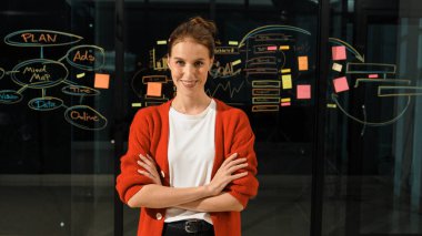 Profesyonel genç yönetici, silah zoruyla kameraya bakıyor. Cam duvarın önünde zihin haritası ve yapışkan notlarla dikilen yetenekli bir işçi. İzleme