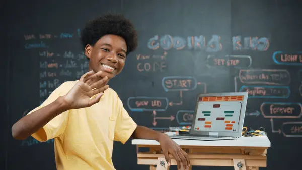 聪明的非洲男孩使用笔记本电脑对程序进行编码 然后转身挥手 在Stem技术教室的黑板上设置漂亮的高中精密编码工程提示或程序系统 — 图库照片