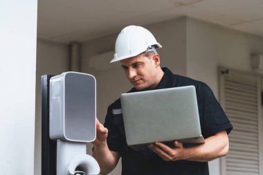 Evde EV şarj istasyonu kurulumu üzerinde çalışan nitelikli teknisyen, EV için dizüstü bilgisayarla şarj sisteminde sorun çıkarma ve yapılandırma ayarları yapıyor. Eşzamanlama