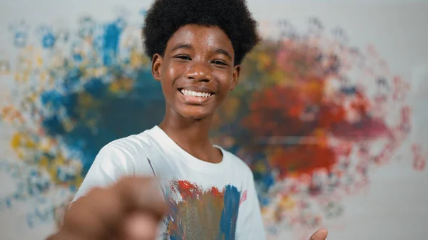 African boy standing at colorful stained wall while holding brush and point at camera. Happy highschool student wearing white shirt with stain color and look at camera with confident. Edification.