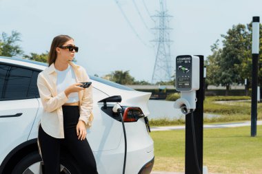 Genç bir kadın EV otomobil şarj istasyonu Green City Park 'ta elektrik ödemek için akıllı telefonu kullanıyor. Modern çevre ve EV araçlı sürdürülebilir şehir yaşam tarzı. Yararlı