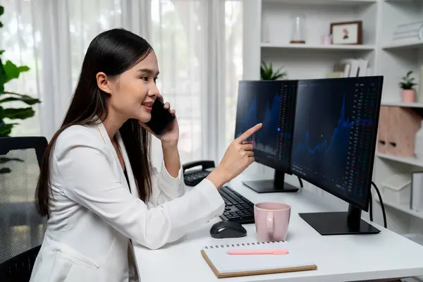 Young beautiful Asian businesswoman calling to broker, pointing dynamic increased exchange stock market graph on pc to invest high profit at modern office, investing high profit in wealth. Stratagem.