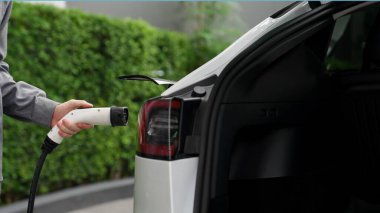 Genç adam yeşil sürdürülebilir şehir açık hava yaz bahçesinde EV elektrikli otomobilini yeniden şarj ediyor kentsel sürdürülebilirlikte gençlerin yaşam tarzı yeşil temiz şarj edilebilir enerji elektrikli araç iç organları