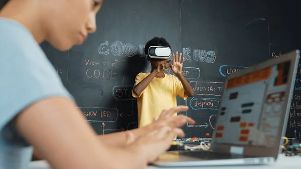 Closeup of school girl hand coding program while african student using VR interact with metaworld. Smart boy wearing virtual reality glasses and playing video games at innovative class. Edification.