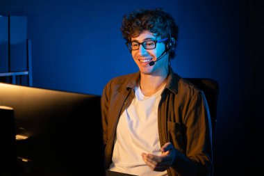 Genç yaratıcı iş adamı, bilgisayar ekranında dinleyicilerle iletişim kuruyor ya da takım üyesiyle görüşüyor. Neon Light Modern Ev Ofisi 'nde kulaklık takıyor. Gece vakti çalışma kavramı. Toplayıcı.