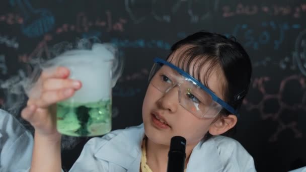 在实验室里 年轻女孩坐在黑板前 用生化理论 拿着绿色化学溶液仔细检查 做实验的年轻学生的衣服 — 图库视频影像