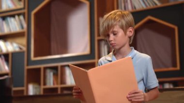 Çekici, beyaz, zeki çocuk kütüphanede kitap okuyor ve bir yığın kitap yerleştirilmiş. Akıllı çocuk bir sayfayı çevirirken okumaya, romandan veya kitaptan öğrenmeye odaklanıyor. Kendi kendini eğitme. Etkinlik.