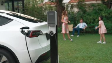 Mutlu ve neşeli bir ailenin arka planında ev şarj istasyonundan elektrikli araç şarj ediliyor. EV arabası daha iyi bir gelecek için alternatif ve sürdürülebilir enerji kullanıyor.