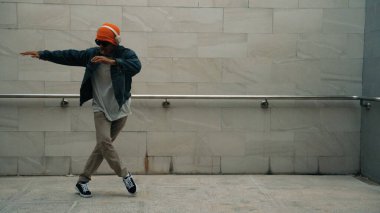 Asyalı akıllı hipster, kulaklık ve şık kıyafetler giyip hip hop şarkıları dinlerken gri arka planlı enerjik break dans gösterisi yapıyor. Açık hava sporu 2024. Hip hop hareketi. Yumuşak bir şekilde..