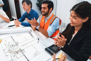 Çeşitli inşaat mühendisi ve müvekkil grupları, mimari proje üzerinde başarılı bir anlaşma sağlandıktan sonra heyecanlanıp alkışlanıyorlar, inşaat planını gözden geçiriyorlar ve toplantı masasında planlarını gözden geçiriyorlar.