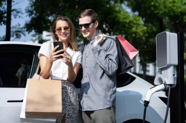 Genç çift yazın yeşil sürdürülebilir şehir bahçesinde EV elektrikli araba ile seyahat ederken, yeşil temiz yeniden şarj edilebilir elektrikli araç iç organları ile kentsel sürdürülebilirlik yaşam tarzını gösteriyor.