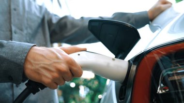 Genç adam yeşil sürdürülebilir şehir açık hava yaz bahçesinde EV elektrikli otomobilini yeniden şarj ediyor kentsel sürdürülebilirlikte gençlerin yaşam tarzı yeşil temiz şarj edilebilir enerji elektrikli araç iç organları