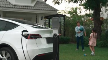 Mutlu ve neşeli bir ailenin arka planında ev şarj istasyonundan elektrikli araç şarj ediliyor. EV arabası daha iyi bir gelecek için alternatif ve sürdürülebilir enerji kullanıyor.