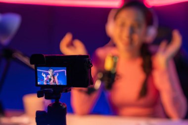 Güzel Asyalı bir kanalın sunucusundaki seçici odak kamera ekranı dinleyiciyle sosyal medyada müzik dinliyor, bulanık arka planda mikrofon kullanarak pastel kulaklık takıyor. Taktik.