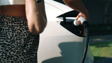 Genç bir kadın yazın yeşil sürdürülebilir şehir bahçesinde EV elektrikli arabasıyla seyahat ediyor. Kentsel sürdürülebilirlik yaşam tarzı yeşil temiz yeniden şarj edilebilir enerji elektrikli BEV araç iç organları