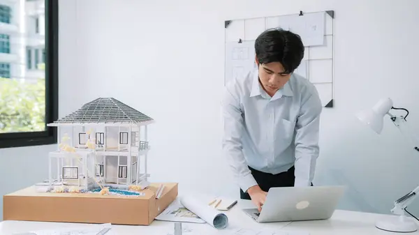 Closeup image of professional architect engineer using laptop analysis data on meeting table with house model and map scatter around at room with blueprint hanged behind. Focus on hand. Immaculate.