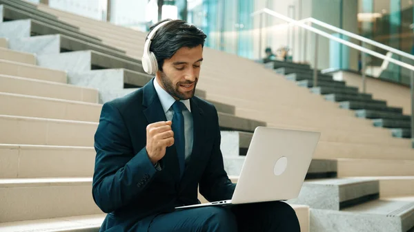 Confident smart business man celebrate successful business idea while listen music. Happy manager project working by using laptop while wearing headphone and suit. Investor sitting at stair. Exultant.