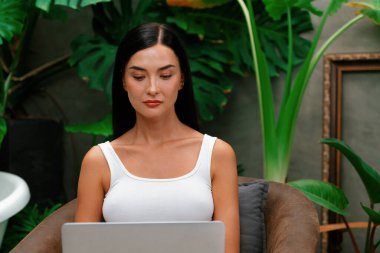 Modern genç kadın, tropikal bitkilerle çevrili, minimalist bir mimari beton stil yaz ekosistem bahçesinde uzaktan çalışıyor ya da dinleniyor. Blithe