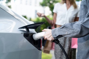 Genç çift yazın yeşil sürdürülebilir şehir bahçesinde EV elektrikli araba ile seyahat ederken, yeşil temiz yeniden şarj edilebilir elektrikli araç iç organları ile kentsel sürdürülebilirlik yaşam tarzını gösteriyor.