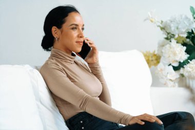 Rahat genç Afrikalı Amerikalı kadın evdeki oturma odasındaki koltukta çok önemli bir cep telefonu kullanıyor.