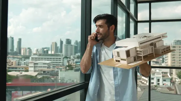 Smart caucasian engineer standing and holding house model while phone calling to manager. Professional manager inspect at architectural model while standing near window. Design concept. Tracery