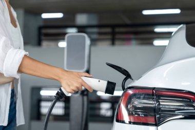 Genç bir kadın EV elektrikli arabasıyla alışveriş merkezinin otoparkına gidiyor şehir merkezinde elektrik iç organlarının yeşil temiz şarj edilebilir enerji ile sürdürülebilirlik yaşam tarzını gösteriyor.