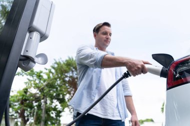 Çevre dostu bilinçli adam EV şarj istasyonundan modern elektrikli aracı şarj ediyor. EV araba teknolojisi gelecekteki sürdürülebilirlik için alternatif ulaşım aracı olarak kullanılıyor. Eşzamanlama