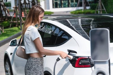 Genç bir kadın yazın yeşil sürdürülebilir şehir bahçesinde EV elektrikli arabasıyla seyahat ediyor. Kentsel sürdürülebilirlik yaşam tarzı yeşil temiz yeniden şarj edilebilir enerji elektrikli BEV araç iç organları