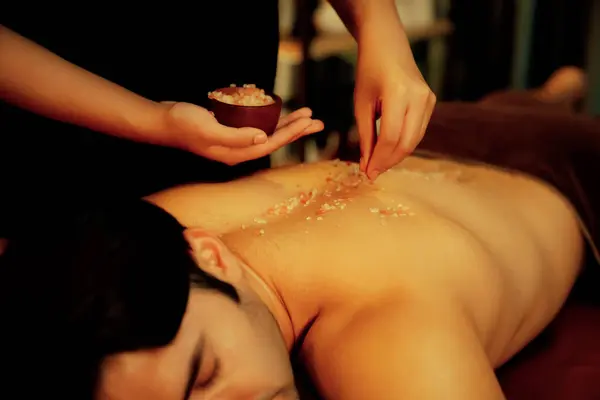 Man customer having exfoliation treatment in luxury spa salon with warmth candle light ambient. Salt scrub beauty treatment in Health spa body scrub. Quiescent