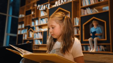 Genç, zeki, beyaz bir kız kütüphanede otururken kitap okuyor. Zeki çocuk öğrenme, okuma, kütüphanede kitap açma. Etkileyici çocuk geçmişi bulanık sayfayı çeviriyor. Etkinlik.