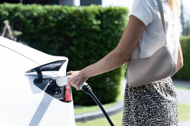 Genç bir kadın yazın yeşil sürdürülebilir şehir bahçesinde EV elektrikli arabasıyla seyahat ediyor. Kentsel sürdürülebilirlik yaşam tarzı yeşil temiz yeniden şarj edilebilir enerji elektrikli BEV araç iç organları