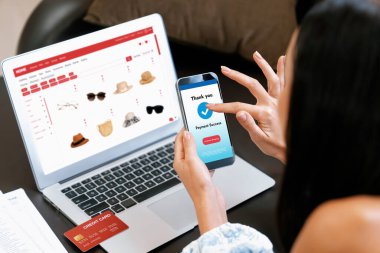 Genç bir kadın dizüstü bilgisayarla ürün sipariş eder ya da satın alır ve akıllı telefondan işlem ödemesi yapar. İnternet bankacılığı vasıtasıyla internet üzerinden kredi kartıyla online alışveriş hayatı. Blithe